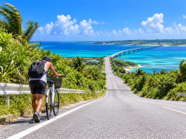 宮古島の橋へ続く上り坂で自転車を押す旅行者